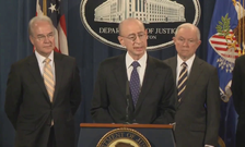 IG Levinson stands at a podium with a Department of Justice logo, on a stage with other officials